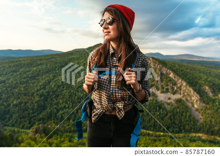 Portrait of a traveler in tourist gear. A young tourist with a backpack admires the sunset from the top of the mountain. Tourist woman with a backpack on a mountain background 85787160