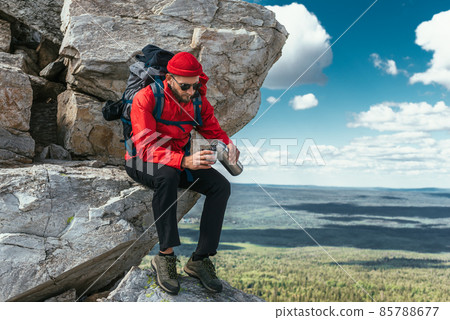 A bearded traveler with a backpack on top of a mountain. Portrait of a traveler in red clothes and sunglasses. A tourist with a backpack sits against the background of mountains. Copy space 85788677