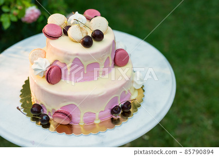 Fruit cake decorated with cherries and French macaroni served on a marble table against the background of a garden with blooming roses Fruit cake decorated with cherries and French macaroni served on a marble table against the background of a garden with blooming roses 85790894