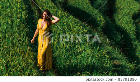 A beautiful girl walks on rice terraces on the island of Bali. A beautiful girl in a yellow dress walks through green rice fields. A young woman walks on rice terraces. Copy space A beautiful girl walks on rice terraces on the island of Bali. A beautiful girl in a yellow dress walks through green rice fields. A young woman walks on rice terraces. Copy space 85791880