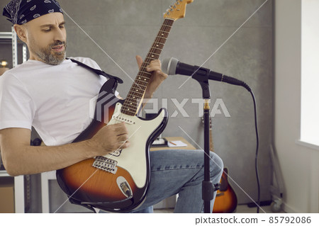 Bearded middle aged man playing his guitar. Male musician rehearsing new song. Hard rock or heavy metal band guitarist in bandana playing riff on electric guitar in modern studio with microphone Bearded middle aged man playing his guitar. Male musician rehearsing new song. Hard rock or heavy metal band guitarist in bandana playing riff on electric guitar in modern studio with microphone 85792086
