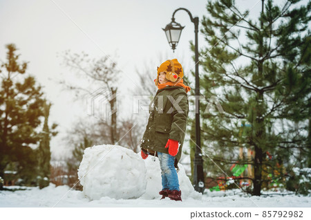 Child with big snow ball while building snowman. Children having fun outdoors on winter holidays vacation and Christmas. Kid playing outdoors on sunny snowy winter day. Happy girl play in sunny day 85792982