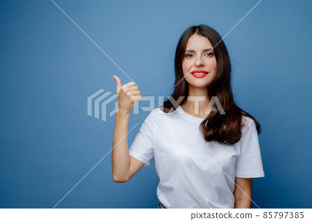 Brunette woman with long hair in a white T-shirt points her thumb to the side. Female portrait isolated against blue solid background. 85797385