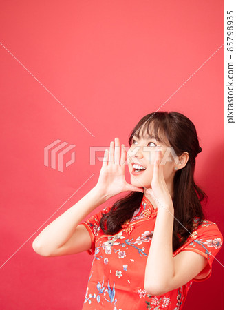 Young asian girl wear a red cheongsam is shouting and holding her hands near her mouth with copyspace on red background. 85798935