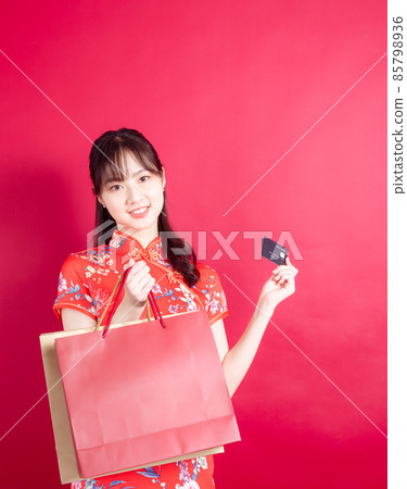 Asian young woman wearing cheongsam qipao dress holding shopping bags and a credit card isolated on red background. Asian young woman wearing cheongsam qipao dress holding shopping bags and a credit card isolated on red background. 85798936