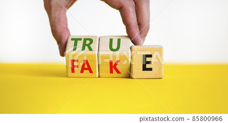 Fake or true symbol. Businessman turns wooden cubes and changes the word fake to true or vice versa. Beautiful yellow table, white background, copy space. Business and fake or true concept. 85800966