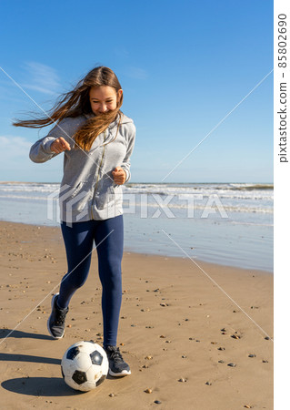 Pre-adolescent Spanish girl running on the beach with her soccer ball while her hair covers her face from the wind. Exercise and fun concept. Pre-adolescent Spanish girl running on the beach with her soccer ball while her hair covers her face from the wind. Exercise and fun concept. 85802690