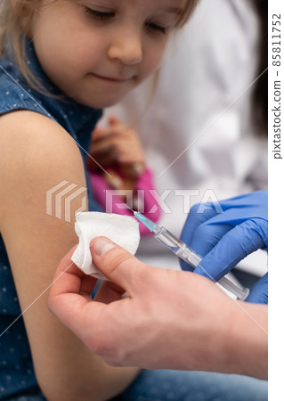 The doctor is getting ready to vaccinate a little girl. A young nurse sits next to the child to make the child feel safe. Preventive vaccine for young children. Close-up frame. The doctor is getting ready to vaccinate a little girl. A young nurse sits next to the child to make the child feel safe. Preventive vaccine for young children. Close-up frame. 85811752