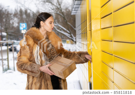 A rich woman in a fur coat operates the touchscreen of a parcel locker. Receive and send parcels automatically in winter. 85811818