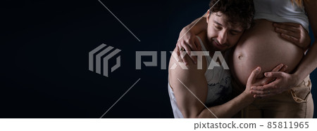 The husband closes his eyes and puts his ear to his wife's pregnancy belly. Hands on the stomach. Ninth month of pregnancy. Blessed state. Panoramic frame. 85811965