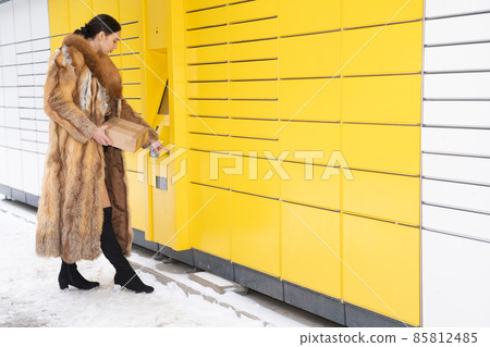 A woman in a fur coat types the code on the numeric keypad of a parcel locker. Receive and send parcels automatically in winter. A woman in a fur coat types the code on the numeric keypad of a parcel locker. Receive and send parcels automatically in winter. 85812485