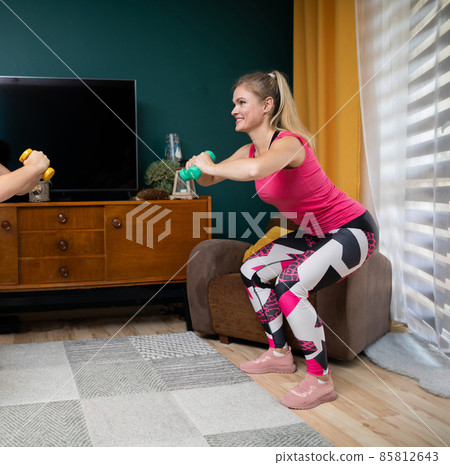 Two girls facing each other are exercising on bent legs and holding dumbbells in their hands. Fitness in the living room. 85812643
