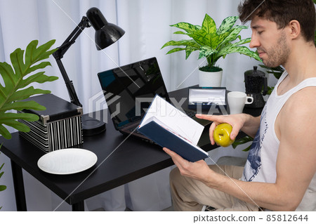 A young worker is holding a green apple in his hand while viewing a book calendar. A domain computer workstation among potted plants with bright lighting. 85812644