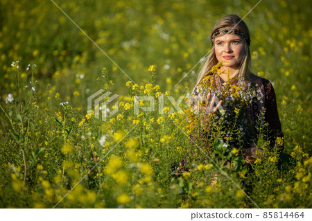 A young woman sits in a field of flowering rapeseed with a bouquet of herbs in her hand. 85814464
