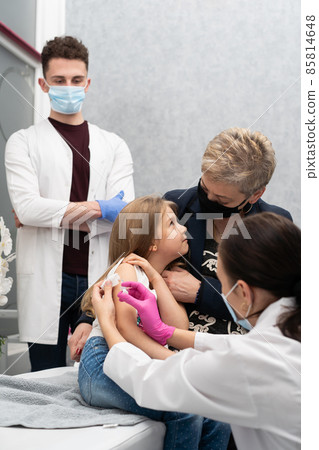 Grandma cheers her granddaughter during the vaccination. A young nurse stabs a needle into the girl's arm. A trainee in the last year of medicine observes the whole situation in the doctor's office Grandma cheers her granddaughter during the vaccination. A young nurse stabs a needle into the girl's arm. A trainee in the last year of medicine observes the whole situation in the doctor's office 85814648