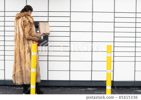 A rich woman in a fur coat operates the touchscreen of a parcel locker. Receive and send parcels automatically in winter. 85815336