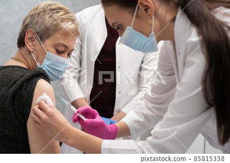 A nurse is making an vaccine to an elderly woman. The COVID vaccine19. A sterile doctor's office in a private clinic. A young medical intern looks at how to properly inject. A nurse is making an vaccine to an elderly woman. The COVID vaccine19. A sterile doctor's office in a private clinic. A young medical intern looks at how to properly inject. 85815338