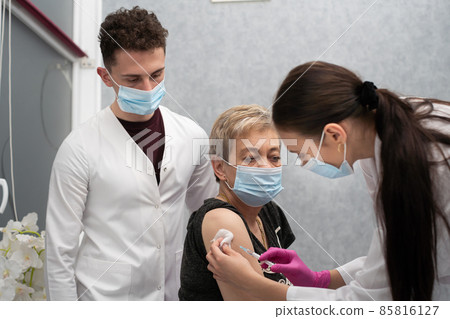 A nurse is making an vaccine to an elderly woman. The COVID vaccine19. A sterile doctor's office in a private clinic. A young medical intern looks at how to properly inject. A nurse is making an vaccine to an elderly woman. The COVID vaccine19. A sterile doctor's office in a private clinic. A young medical intern looks at how to properly inject. 85816127