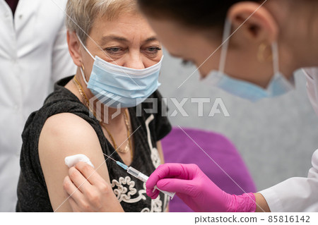 A nurse is making an vaccine to an elderly woman. The COVID vaccine19. A sterile doctor's office in a private clinic. A young medical intern looks at how to properly inject. 85816142