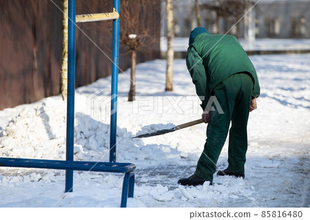 A manual worker scoops snow from the pavement and shovels it onto a pile next to it A manual worker scoops snow from the pavement and shovels it onto a pile next to it 85816480