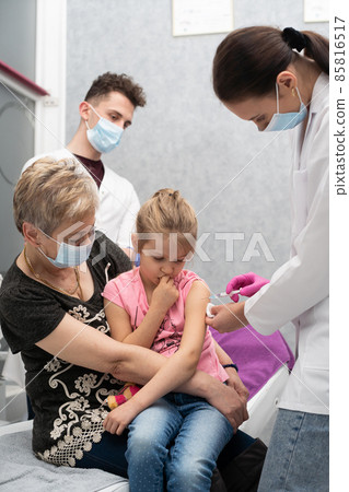 A nurse gives a vaccine to a girl who is sitting on her grandmother's lap because she feels so safe. A trainee in the last year of medicine observes the whole situation in the doctor's office and A nurse gives a vaccine to a girl who is sitting on her grandmother's lap because she feels so safe. A trainee in the last year of medicine observes the whole situation in the doctor's office and 85816517