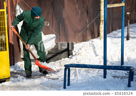 A manual worker scoops snow from the pavement and shovels it onto a pile next to it A manual worker scoops snow from the pavement and shovels it onto a pile next to it 85816524