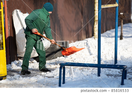 A manual worker scoops snow from the pavement and shovels it onto a pile next to it A manual worker scoops snow from the pavement and shovels it onto a pile next to it 85816533