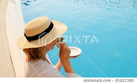 Close up of a woman aged 50-55 in a straw hat drinking from a cup of coffee next to a blue swimming 85822380