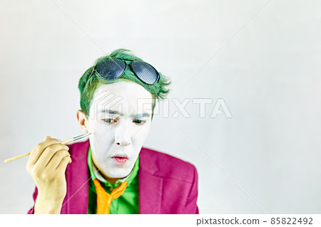 Mim is preparing for performance. Cosplayer guy paints his face with brush. Actor with white makeup and green hair in burgundy suit with yellow tie. Background with copy space for text. 85822492