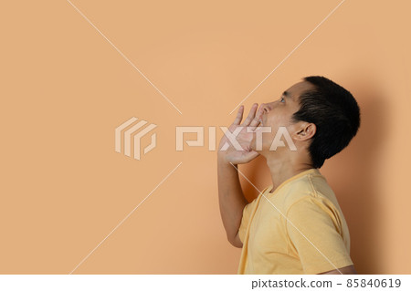 Young man shouting and screaming loud to side with hand on mouth isolated on orange studio background 85840619