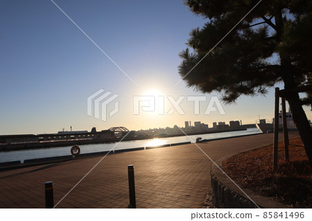 A view of the sea, the harbor, and the building town of the coastal park with the light orange sun, rescue floats and trees, and the glittering road. A view of the sea, the harbor, and the building town of the coastal park with the light orange sun, rescue floats and trees, and the glittering road. 85841496