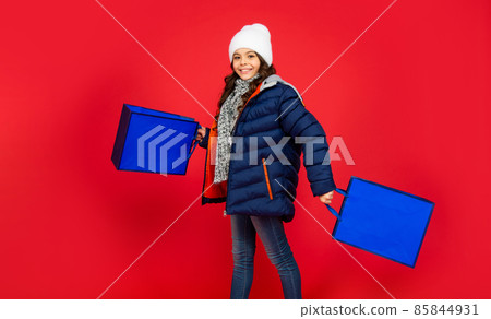 happy kid in puffer jacket and hat. teen girl after shopping on red background. happy kid in puffer jacket and hat. teen girl after shopping on red background. 85844931