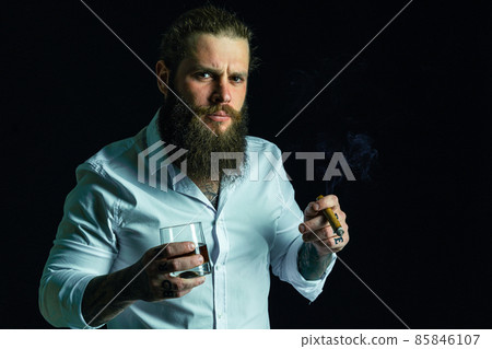 hipster bearded man in white shirt holds cigar and glass of whiskey, looking at camera. Studio shot 85846107