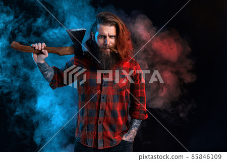 Bearded lumberjack. Bearded man with cigar in checkered shirt with long hair holds old axe in smoke 85846109