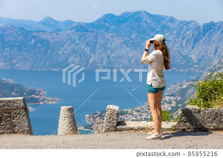 Young woman looking at Kotor bay in Montenegro through binoculars at view spot on serpantine road Young woman looking at Kotor bay in Montenegro through binoculars at view spot on serpantine road 85855216