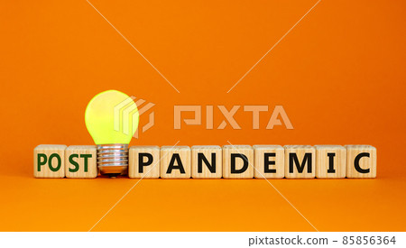 Post-pandemic time. Wooden cubes with word 'post-pandemic'. Yellow light bulb. Beautiful orange background. Business and covid-19 post-pandemic concept. Copy space. 85856364