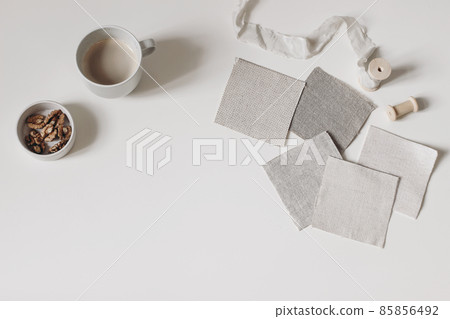 Artistic craft still life. Beige fabric linen swatches and silk ribbon isolated on white table background. Cup of coffee, bowl with walnuts. Neutral mood board project flat lay, top. Fabric samples. 85856492