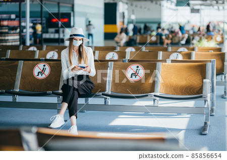 Woman tourist with mask with flight tickets and sitting in a socially distanced chair at the airport during the coronavirus epidemic. Concept of new normality of air transportation due to pandemic. Woman tourist with mask with flight tickets and sitting in a socially distanced chair at the airport during the coronavirus epidemic. Concept of new normality of air transportation due to pandemic. 85856654