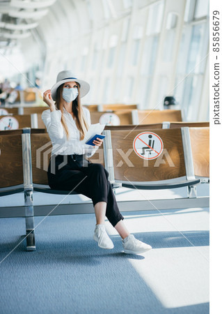 Tourist in mask sitting in airport terminal lounge on empty marked chairs under new normal and covid social distance regulations. Woman sitting in airport lounge and looking at her passport and travel Tourist in mask sitting in airport terminal lounge on empty marked chairs under new normal and covid social distance regulations. Woman sitting in airport lounge and looking at her passport and travel 85856679