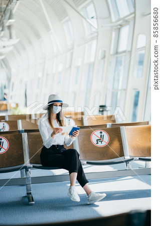 Young girl, business woman standing by the window in a modern airport holding tickets and a phone, looking at the camera. Portrait of a beautiful girl in a mask. Business trip by plane. Young girl, business woman standing by the window in a modern airport holding tickets and a phone, looking at the camera. Portrait of a beautiful girl in a mask. Business trip by plane. 85856680
