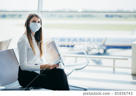 Young girl, business woman standing by the window in a modern airport holding tickets and a phone, looking at the camera. Portrait of a beautiful girl in a mask. Business trip by plane. 85856743