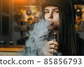 Young pretty woman in cap smoke an electronic cigarette at the vape shop. Hip-hop style. Closeup. 85856793