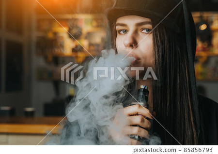 Young pretty woman in cap smoke an electronic cigarette at the vape shop. Hip-hop style. Closeup. 85856793