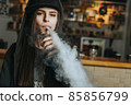 Young pretty woman in cap smoke an electronic cigarette at the vape shop. Hip-hop style. Closeup. 85856799