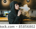 Young pretty woman in cap smoke an electronic cigarette at the vape shop. Hip-hop style. Closeup. 85856802