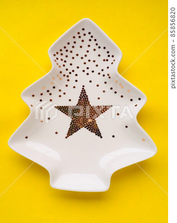 White plate in the shape of a Christmas tree with gold decorations and a star in the middle. Yellow background. New Year or Christmas card. Background for advertising and congratulations. Copy space 85856820