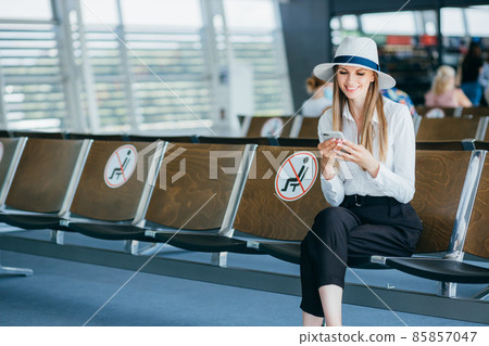 Young woman in a protective face mask sitting in the airport lounge and uses her smartphone. Sends text messages, looks at the camera, checking mails on the Internet, a successful woman leader at work Young woman in a protective face mask sitting in the airport lounge and uses her smartphone. Sends text messages, looks at the camera, checking mails on the Internet, a successful woman leader at work 85857047