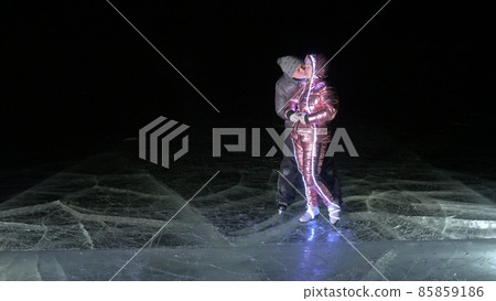 Young loving couple skating at ice rink at night. Man and woman kiss hug have fun learn to skate. Making love on ice. Ice illuminated backlit below. 85859186