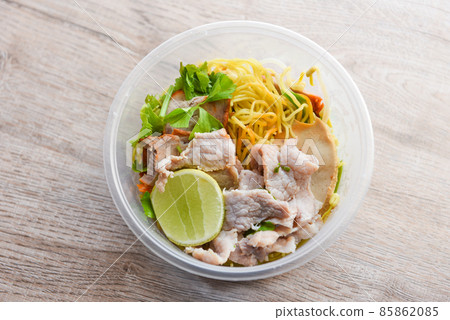 Dry noodles pork with red roasted pork and vegetable in plastic bowl on wooden table, top view Thai and China Asian food yellow noodles lemon lime 85862085
