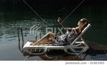 Woman shooting on handheld film gimbal stabilization for smartphone. Girl lie sunbed on pier make selfie. Blogger broadcast video vlogging. 85863993